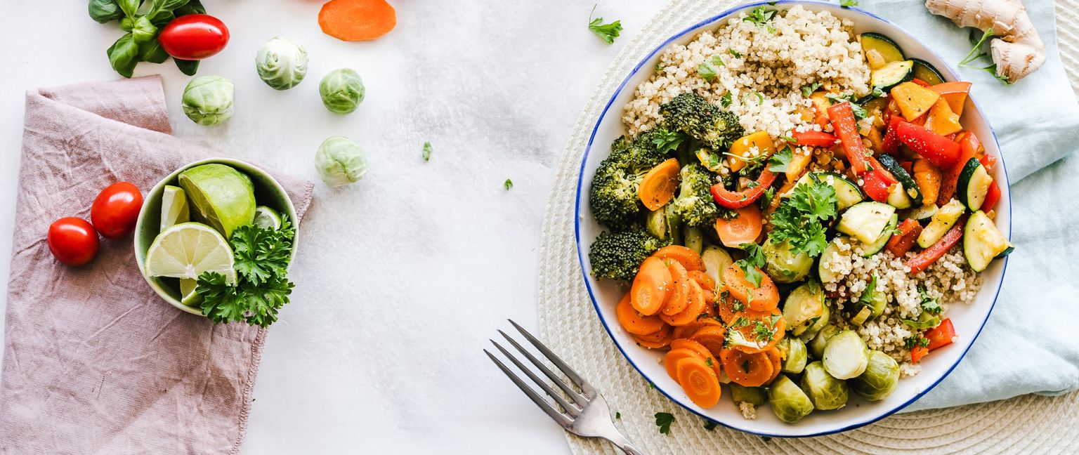 A large bowl of a Mediterranean-style meal, including quinoa, broccoli, carrots, brussels sprouts, zucchini, and red bell pepper. To the side are whole tomatoes, brussels sprouts, and a bowl of sliced lime and parsley.