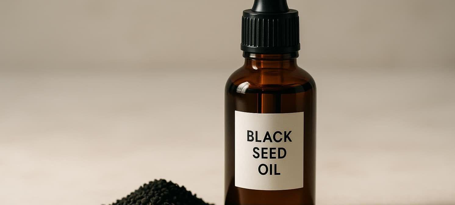 An amber glass bottle of black seed oil with a black dropper top, labeled 'BLACK SEED OIL,' sits beside a pile of black cumin seeds on a light, neutral surface.