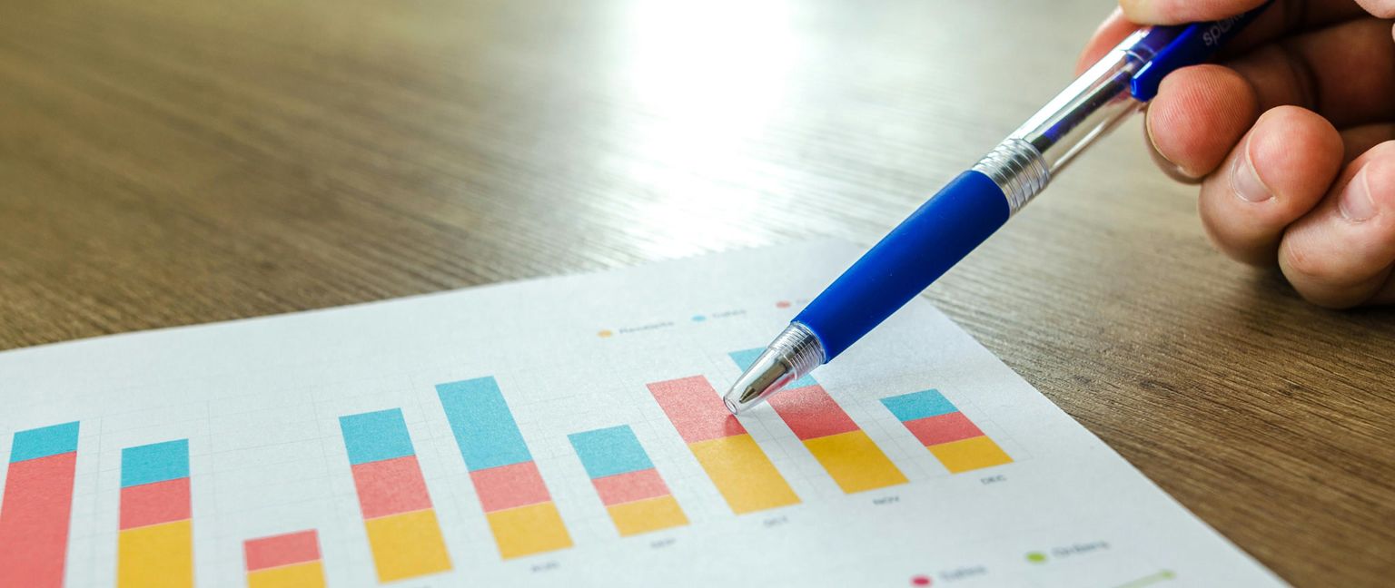 A hand holding a blue pen points at a bar chart with red, yellow, and blue bars on a white sheet of paper.