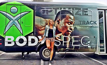 Kim Glass stands smiling in front of a large truck with custom graphics that say 'Body Spec' and feature a large close-up image of a man's face.