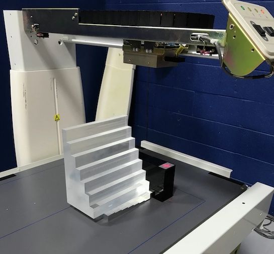 A clear, stepped acrylic phantom rests on the table of a DEXA scanner, likely used for quality assurance testing.