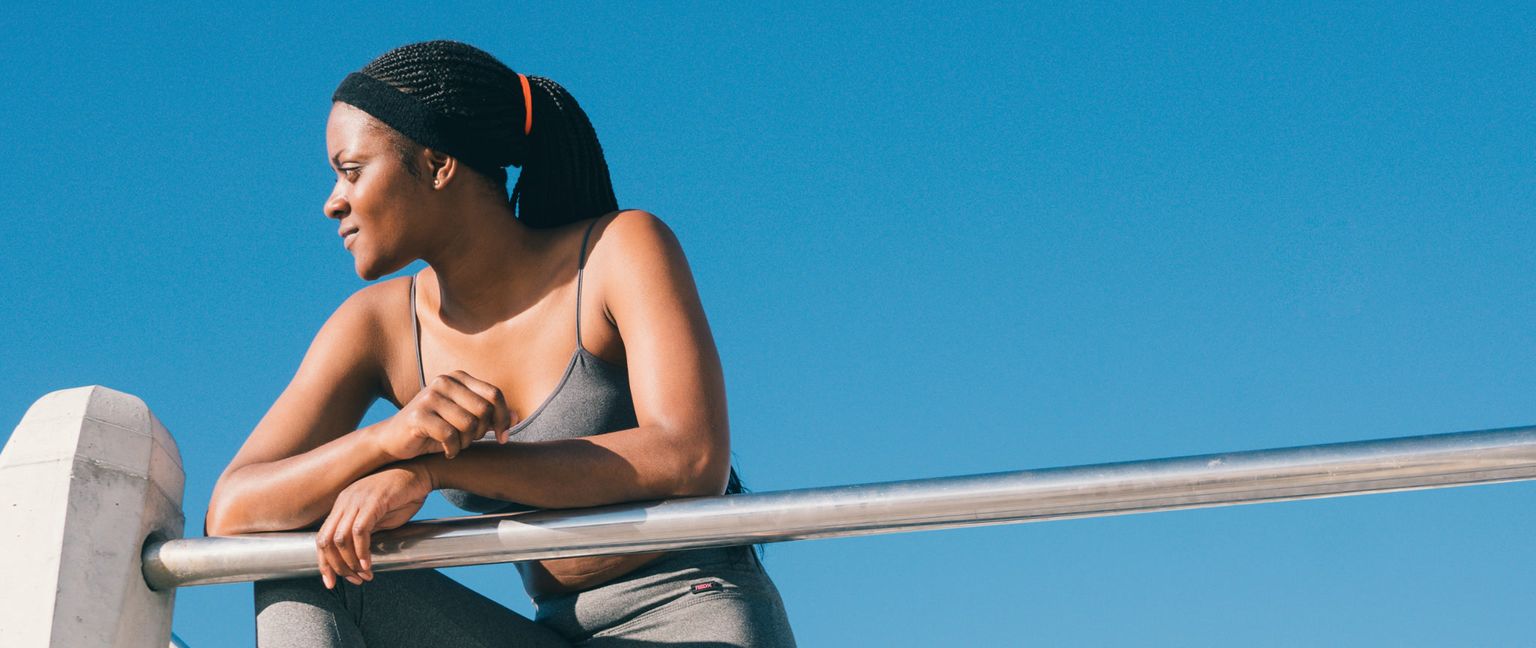 A woman in a grey sports bra and leggings rests her arms on a metal bar and looks away from the camera under a clear blue sky.