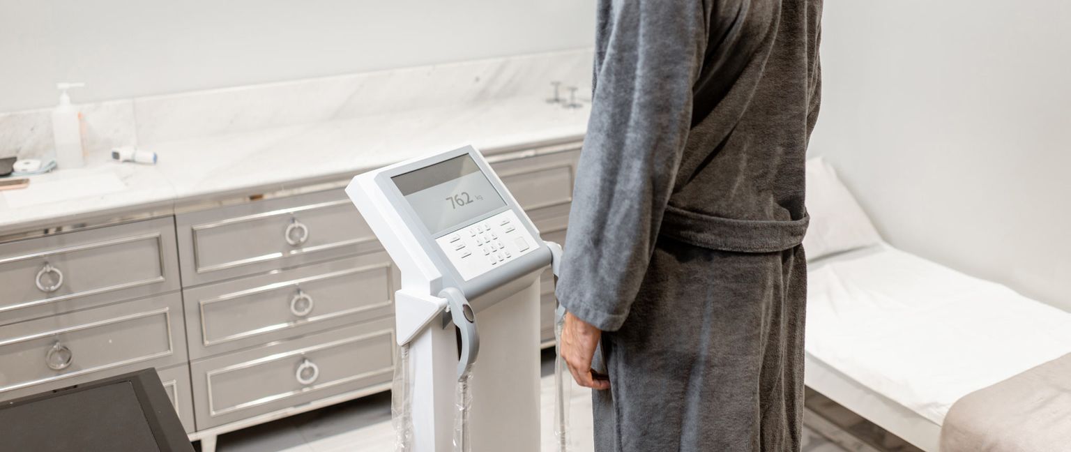 A man in a gray robe stands on an InBody machine, which displays a weight of 76.2 kg. In the background, there is a gray cabinet, a counter with medical supplies, and a bed.