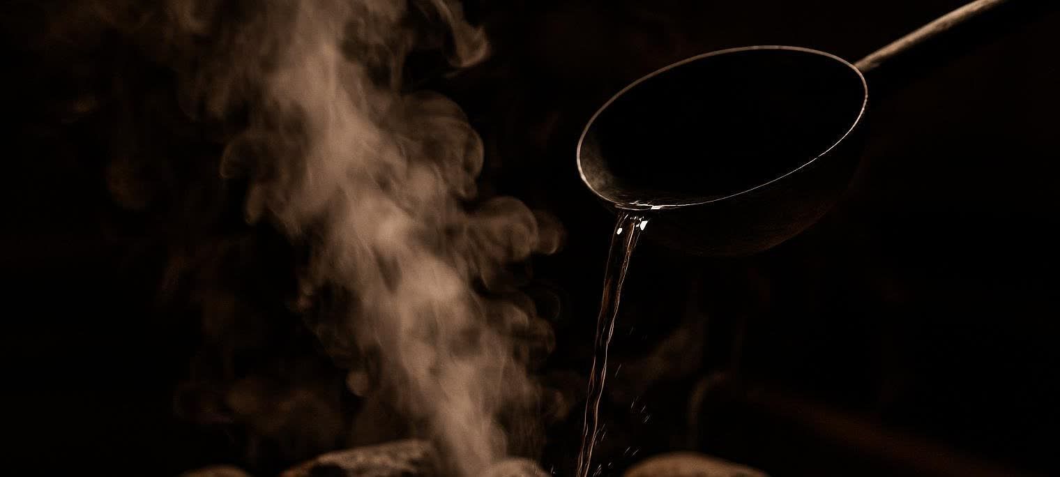 Close-up of a dark ladle pouring water onto hot rocks, creating a plume of steam in a dimly lit sauna.