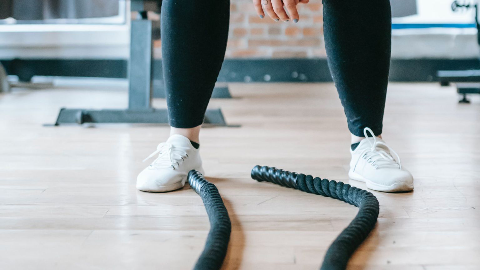 A close-up view of a person's legs standing in a gym with black battle ropes on the floor in front of them.