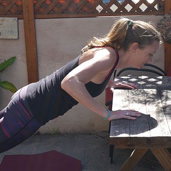 A woman in athletic wear leans on a wooden table outside in a pushup position.