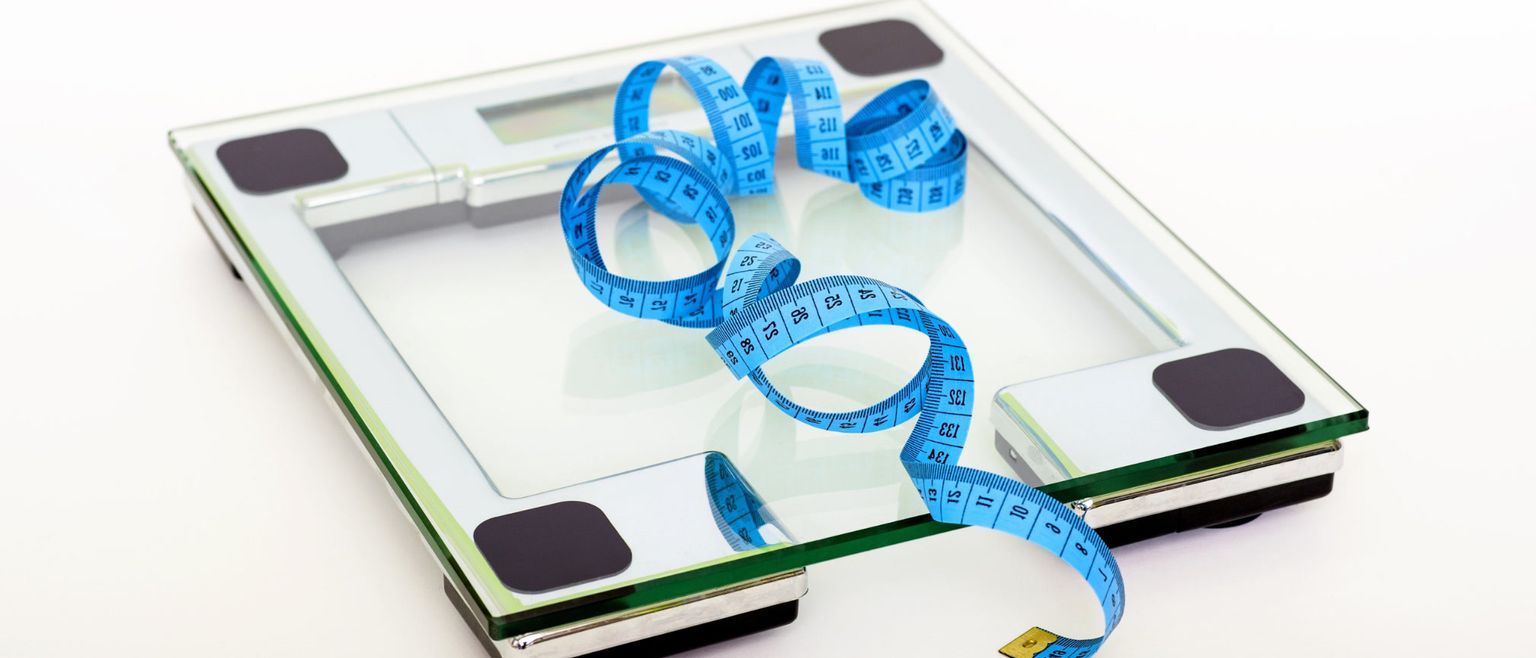 A digital bathroom scale with a blue measuring tape draped across it, on a white background.