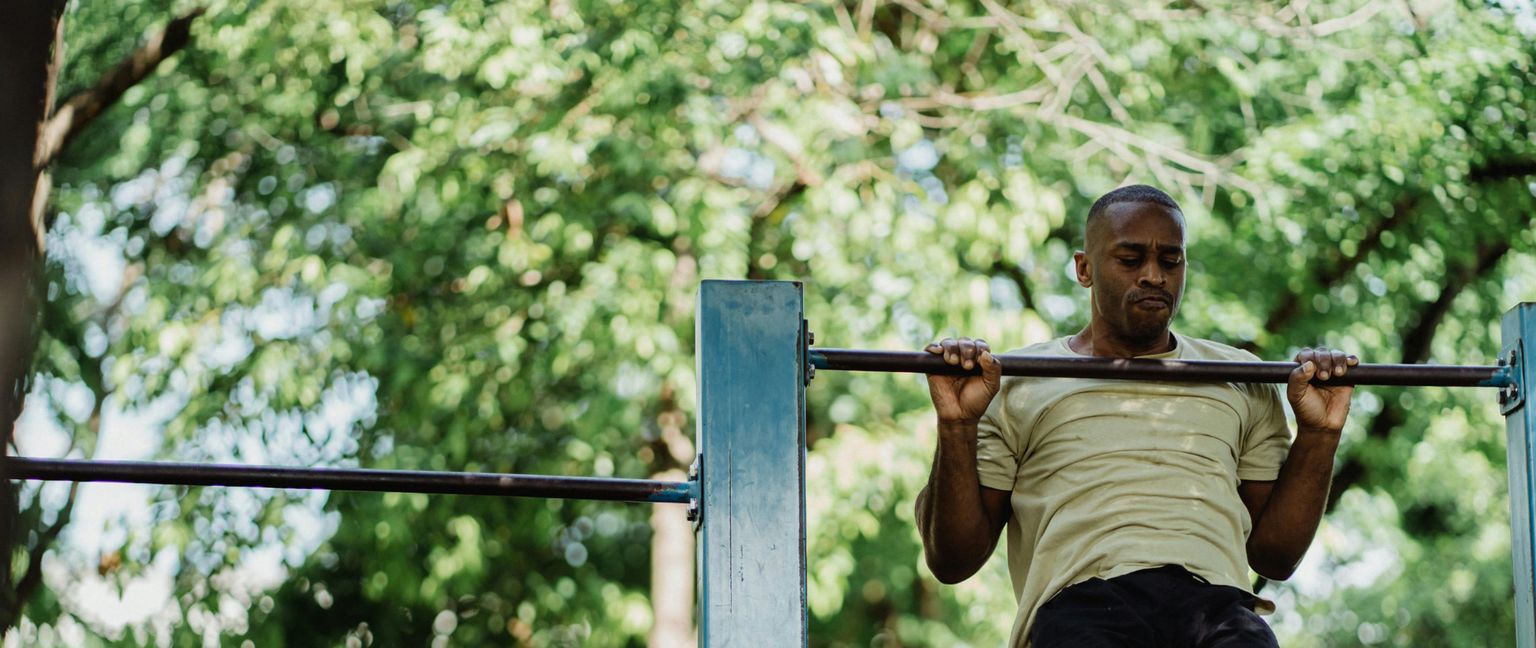 A man in a light green shirt and dark shorts doing a pullup on a metal bar in a park.