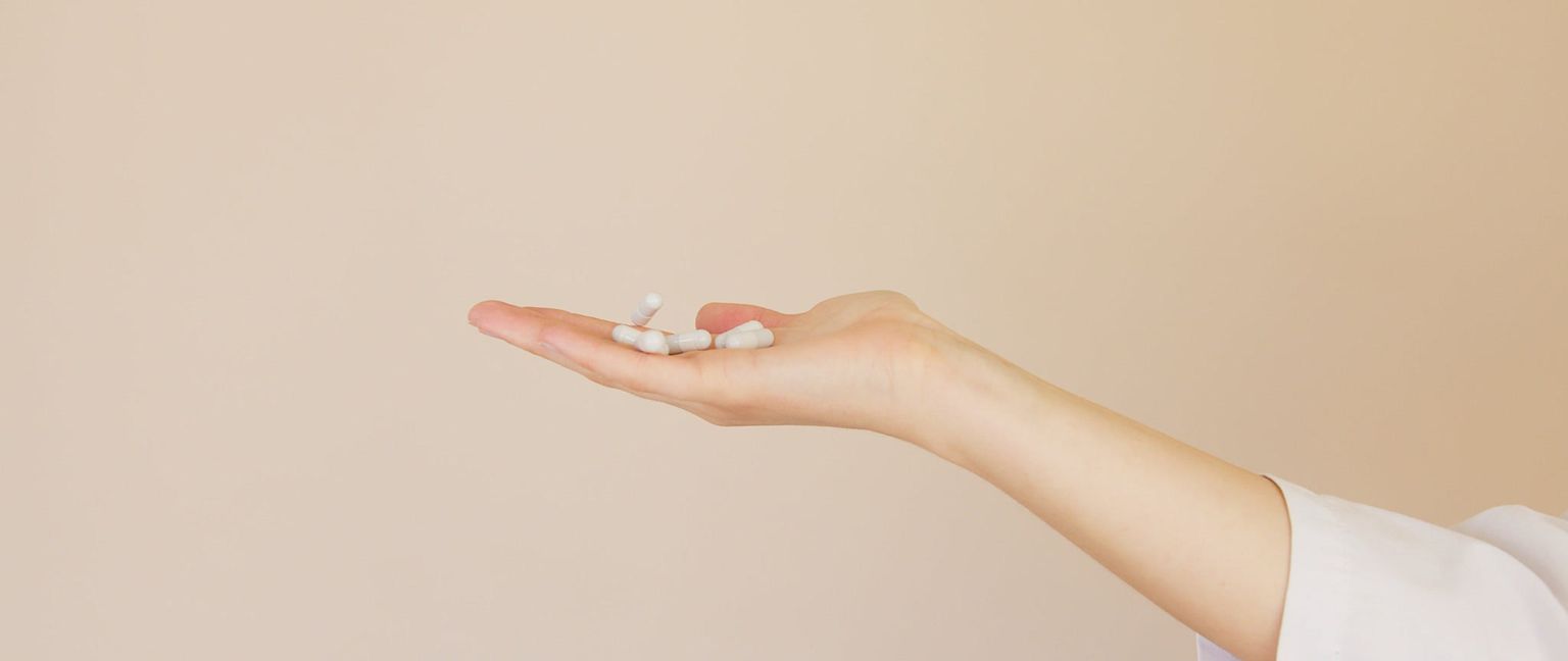 A person's hand holds several white pills or capsules against a tan background.