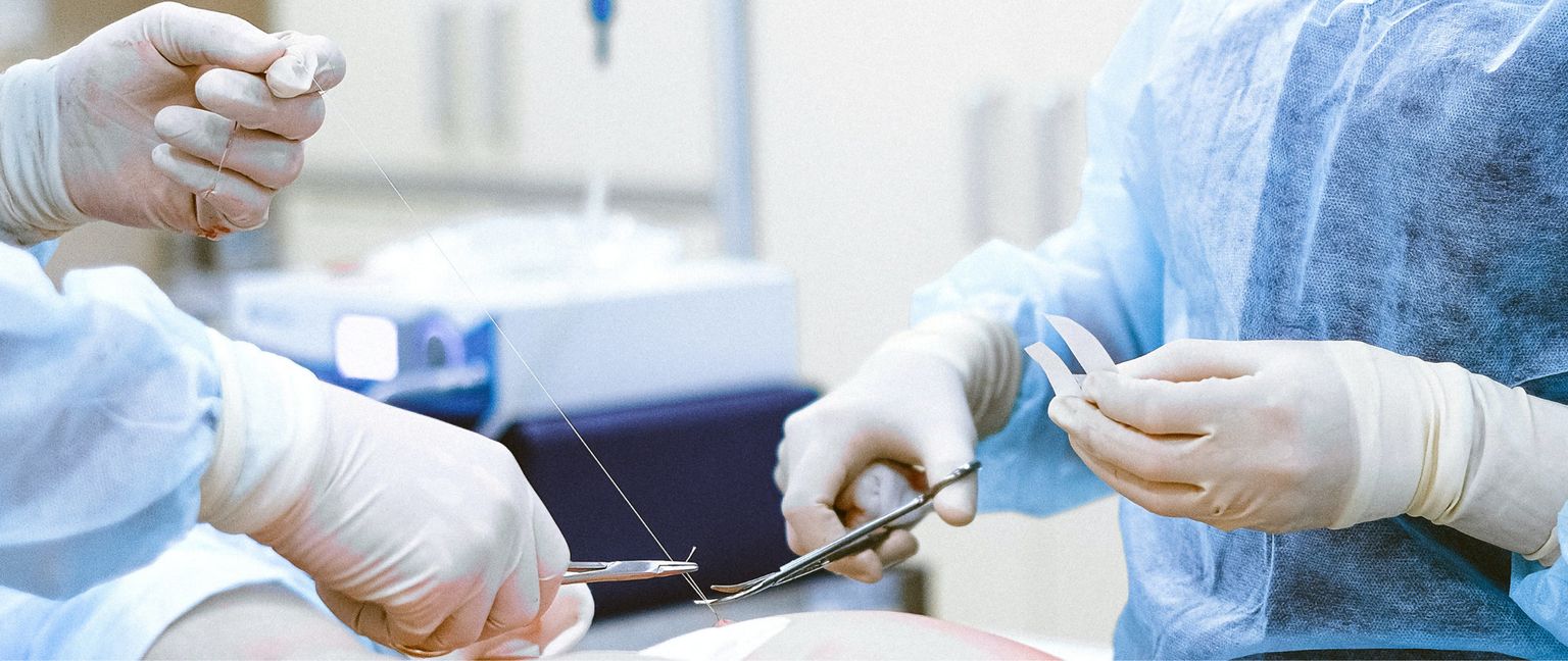 Close up of two surgeons in a sterile operating room wearing disposable gowns and latex gloves, one is suturing a wound with forceps and silk thread, and the other is holding pieces of gauze pads with forceps.