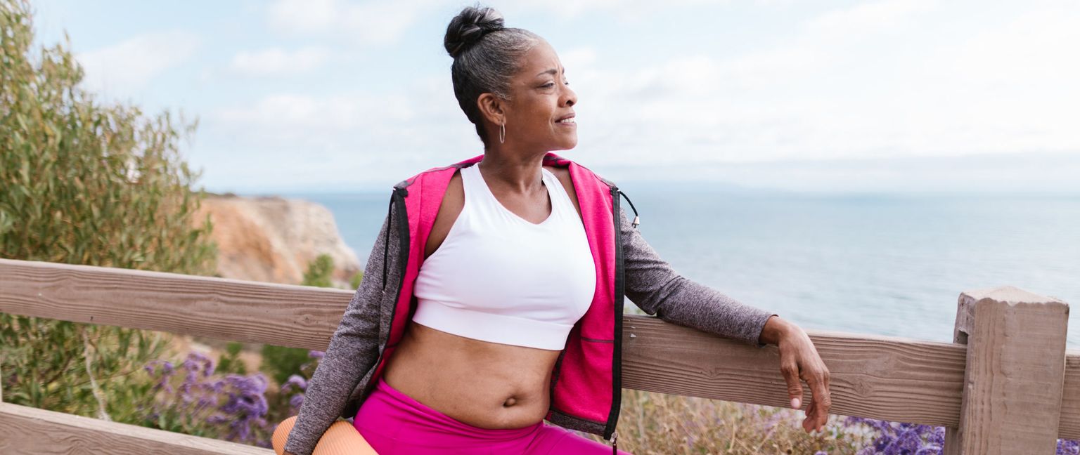 A mature woman with gray hair tied back in a bun is resting her arm on a wooden fence after hiking. She is wearing a white sports bra, pink leggings, and a partially unzipped pink jacket. The ocean is visible in the background.