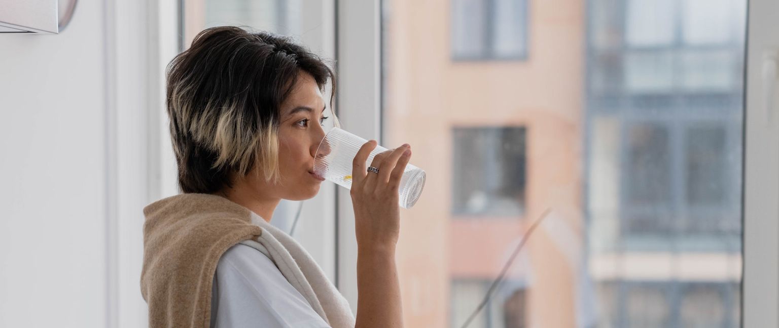 A person is drinking water from a glass while standing near a window.