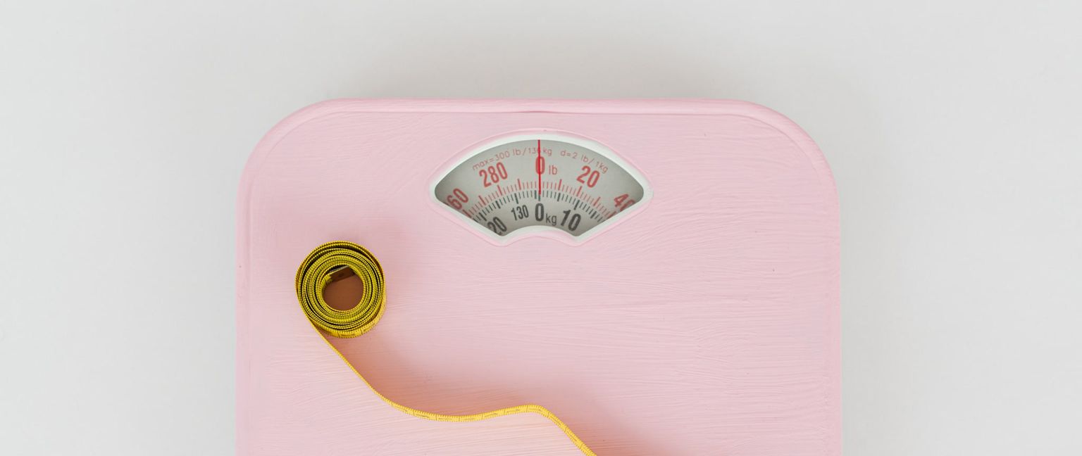 A close-up of a pink scale. A coiled yellow measuring tape is placed on the left side of the scale, with the tape unfurling to the right.