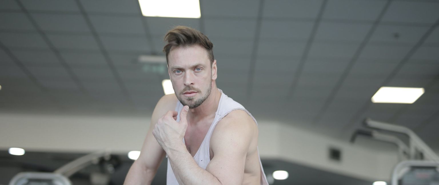 A man in a white tank top in a gym, looking at the camera with his hand on his chin as if deep in thought.