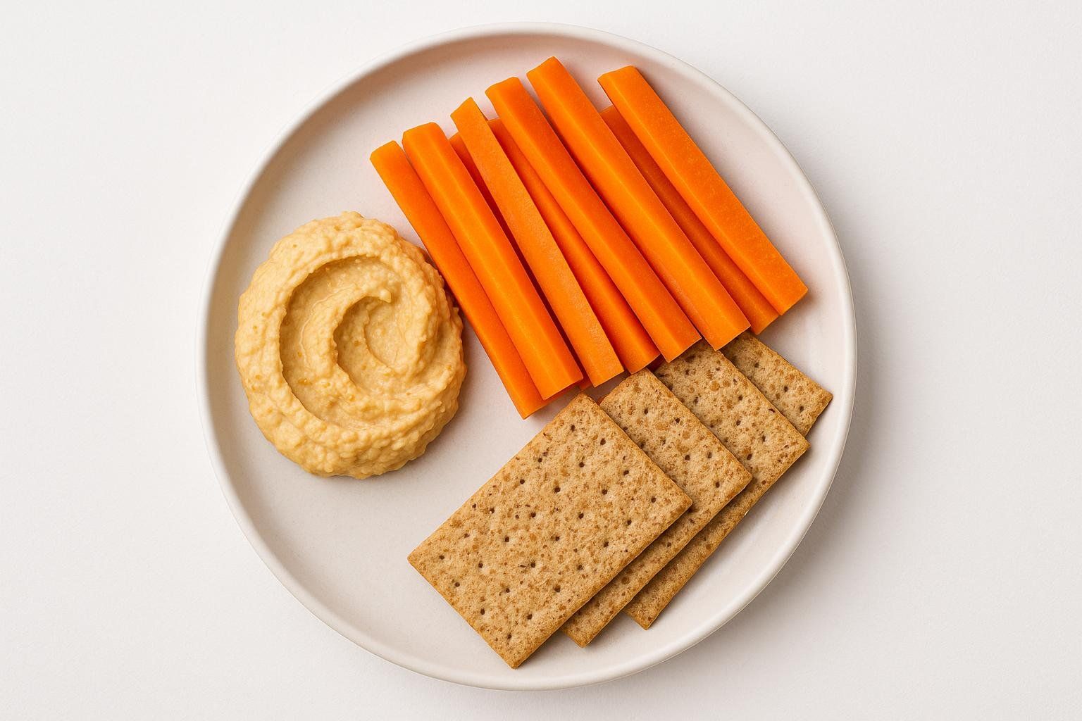 A white plate with a swirl of hummus, neatly arranged carrot sticks, and rectangular crackers. The items are spread across the plate, suggesting a healthy snack.