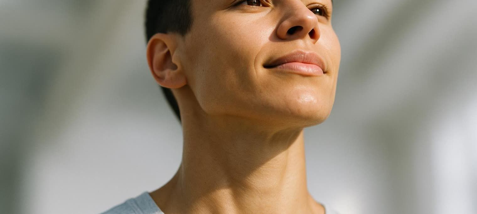 Close-up front-facing photo of a person with short hair, an uplifted face and defined jawline, looking towards the top right with a slight smile. The person's skin is clear and smooth, and dappled with sunlight.