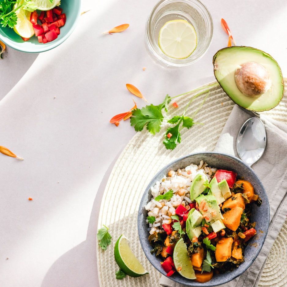 An overhead view of a healthy Buddha bowl filled with rice, sweet potato chunks, avocado slices, red bell peppers, and cilantro. A glass of water with a lime slice, a half avocado, and a small bowl of chopped red peppers and lime wedges are also on the table.