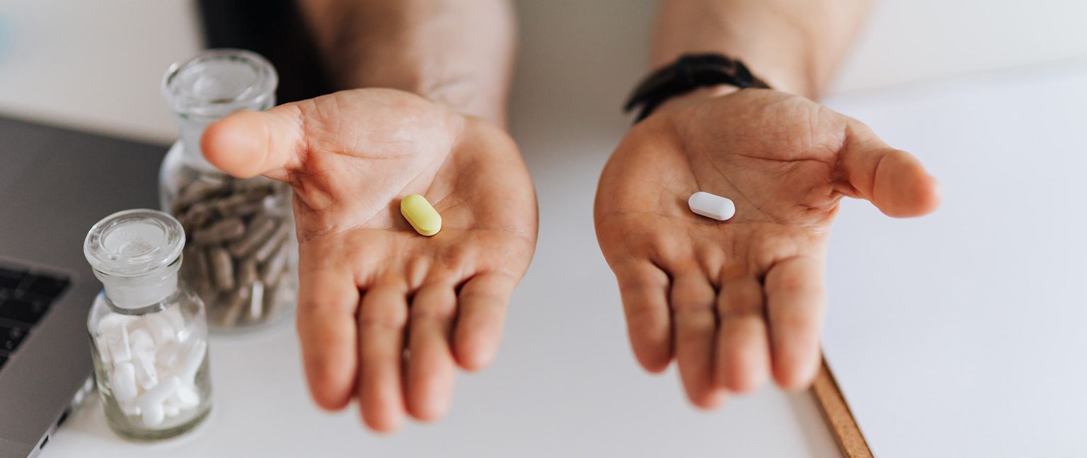A person is holding up two different color pills for comparison in their hands. One pill is white and being held up in their left hand, while the other pill is a light yellowish color and being held up in their right hand. Two glass bottles are visible in the background.