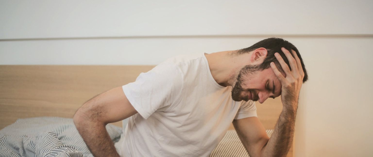 A man in a white t-shirt is sitting up in bed, holding his head in his hand with a pained expression, indicating a headache.