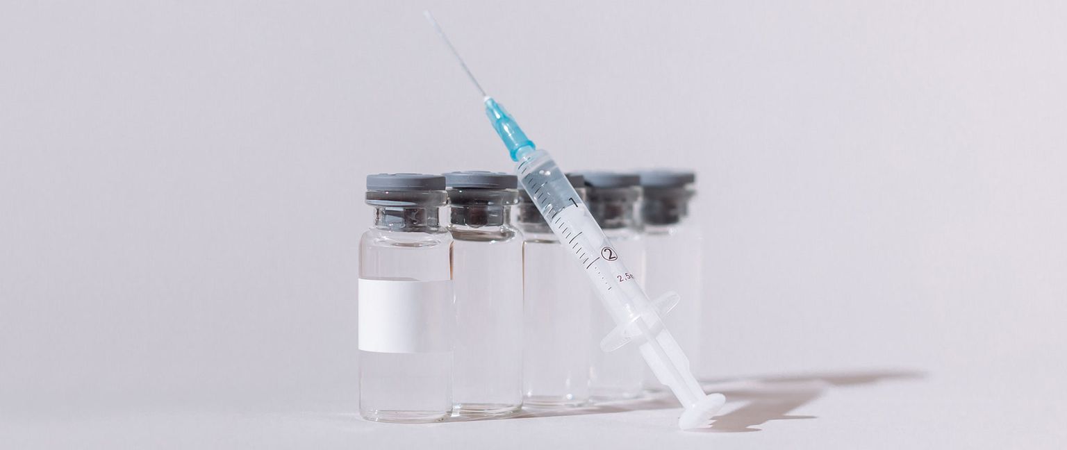 Five clear vaccine vials stand upright with a syringe leaning against the front vial.