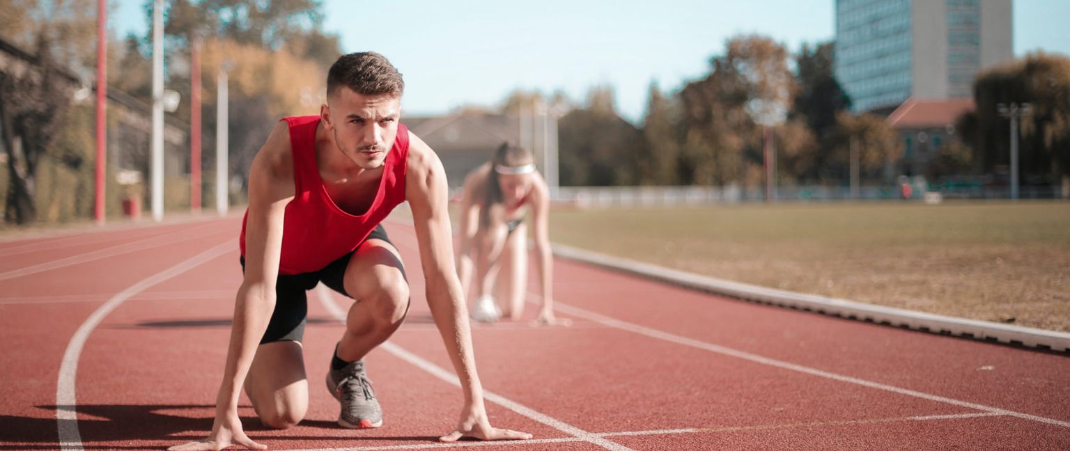 A man in a red tank top and black shorts is in the starting position on a running track. Another runner is visible in the background.