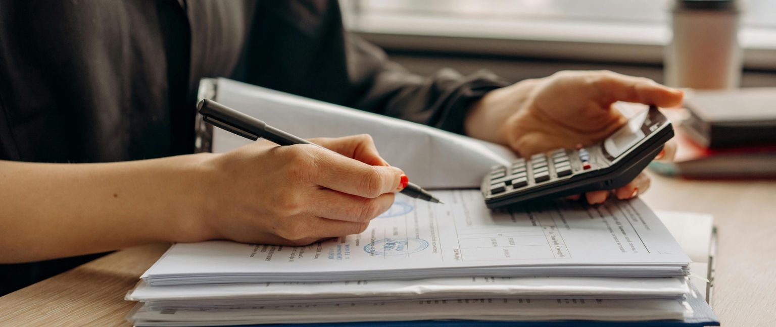 A person's hands are visible. One hand holds a pen to write on a document while the other hand is poised over a calculator. There is a stack of papers on the table.