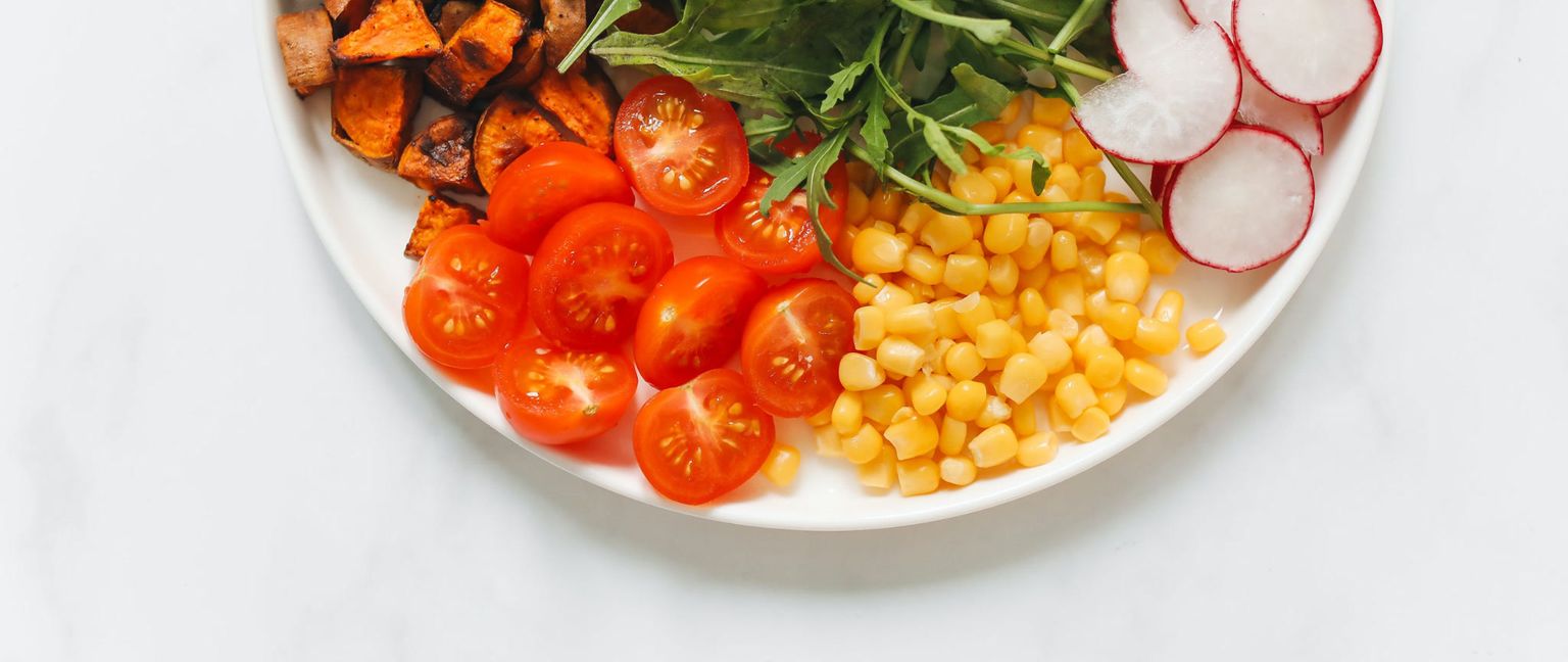 Close-up of a white plate filled with a variety of vegetables including roasted sweet potatoes, halved cherry tomatoes, corn kernels, arugula, and sliced radishes.