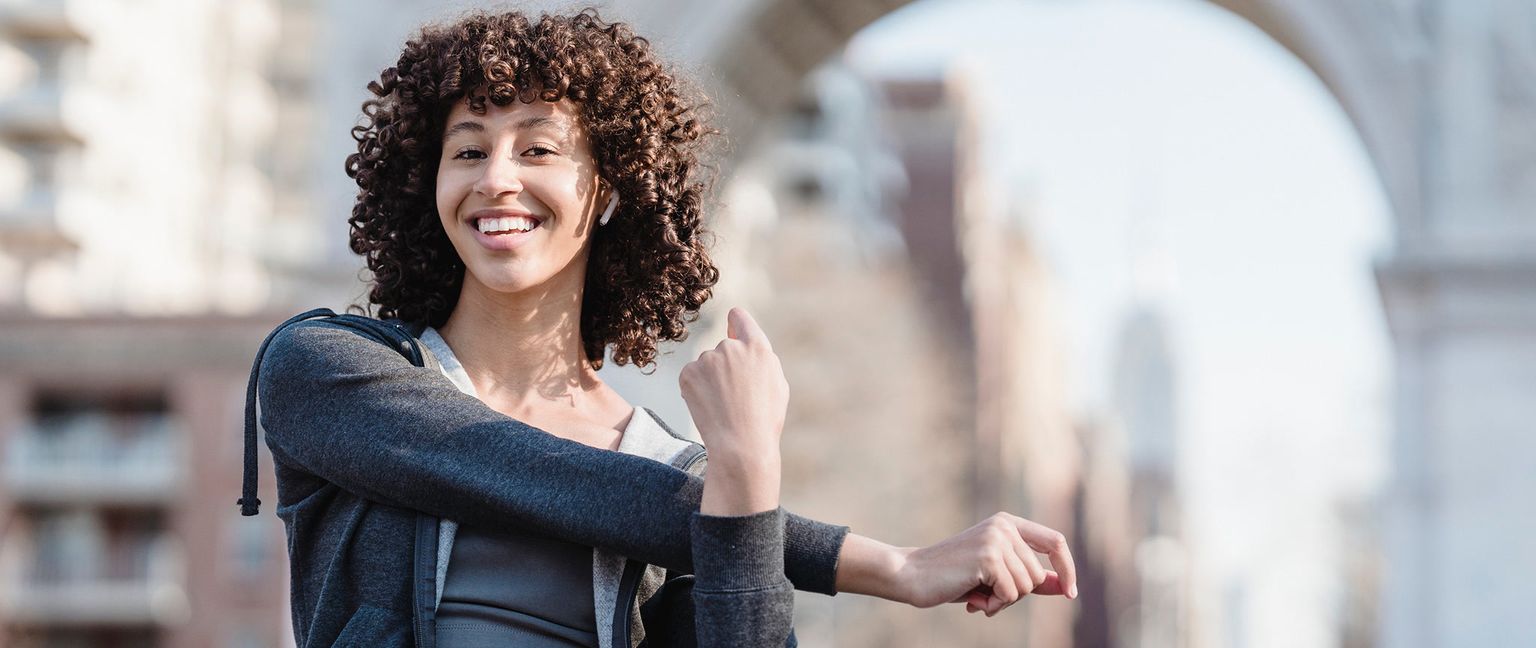 A woman with curly hair stretches her arm across her chest, smiling at the camera outdoors. She is wearing a grey hooded jacket and has wireless earbuds in her ears. A blurry building or archway is visible in the background.