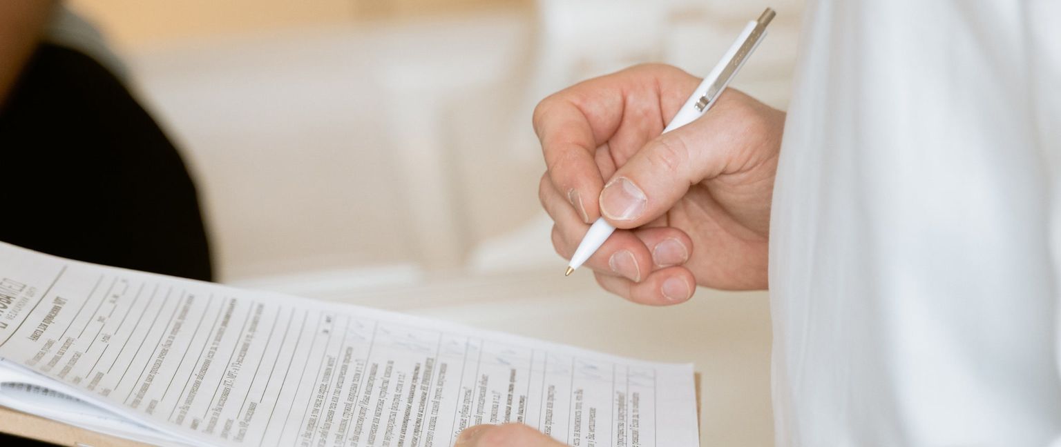 A person's hand is shown holding a white pen and filling out a medical intake form.