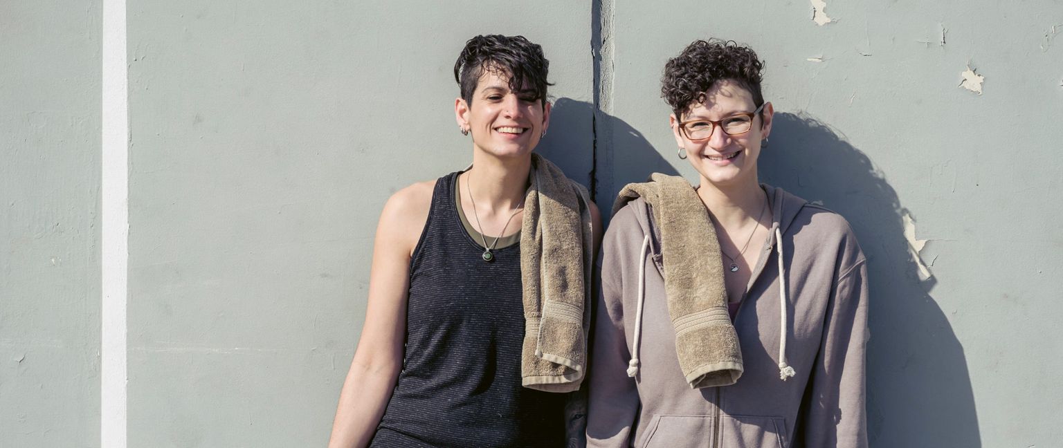 Two women stand outside with towels draped over their shoulders, smiling at the camera.
