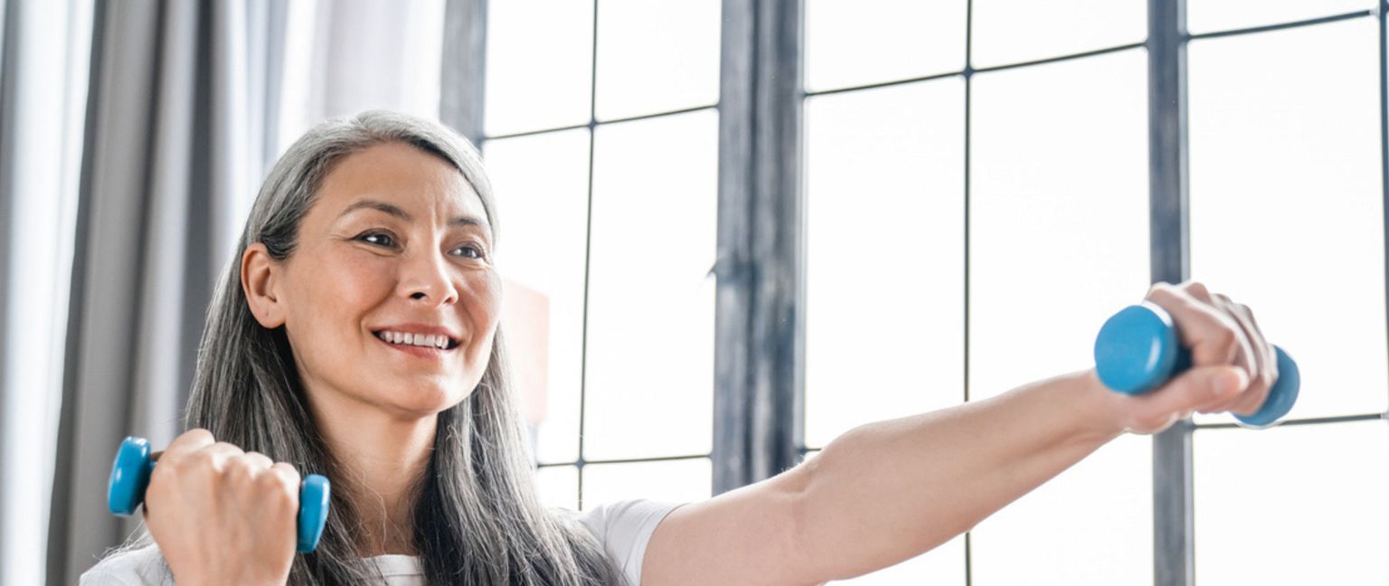 A middle-aged woman with gray hair smiles while holding a small blue dumbbell in each hand.