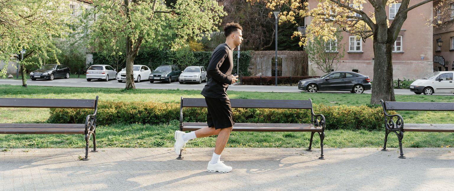A man in a black long sleeve shirt and black shorts runs along a paved path in a park. Several parked cars are visible in the background.