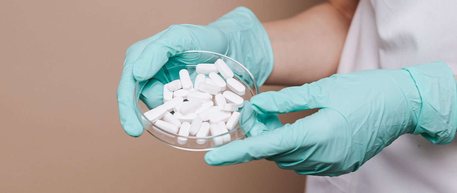 Close-up of gloved hands holding a glass dish filled with white, oblong pills.