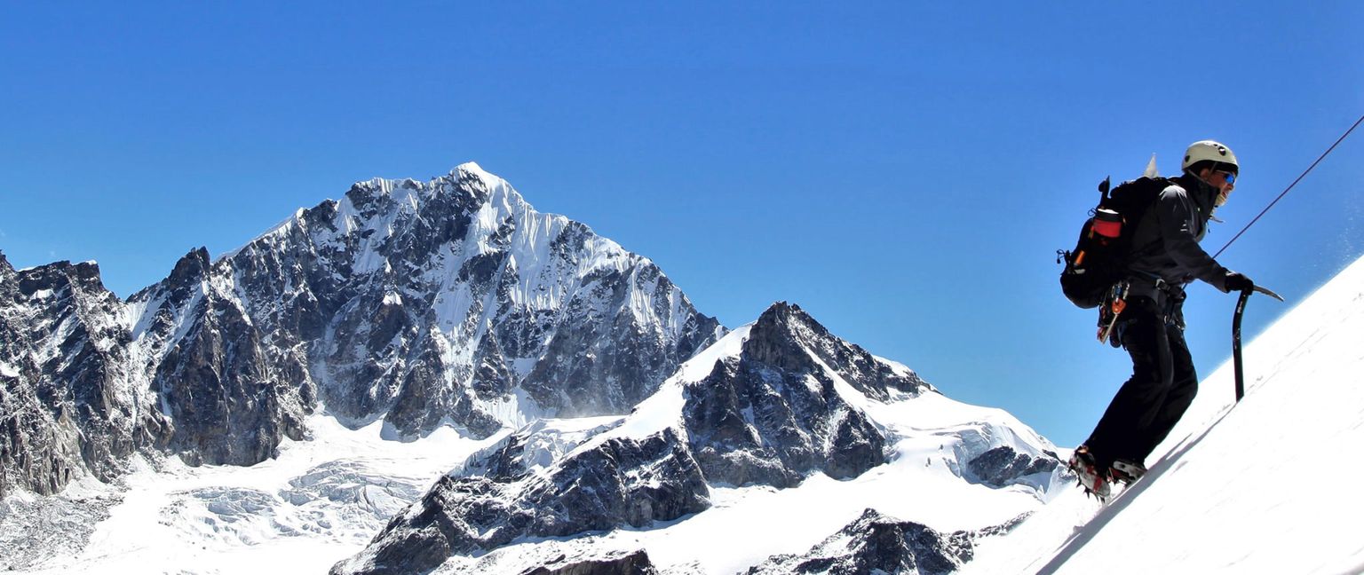 A man in climbing gear ascends a steep, snow-covered mountain slope with an ice axe. Tall, jagged snow-capped mountains are visible in the background under a clear blue sky.