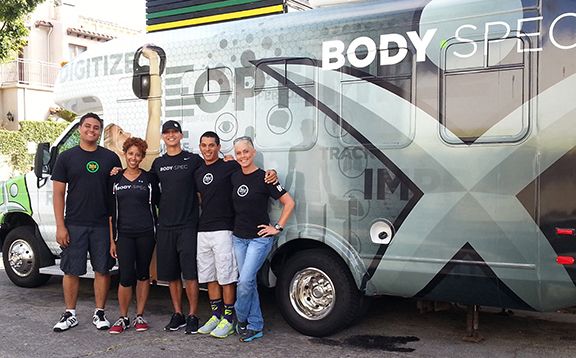 Five people wearing black t-shirts with "BODY SPEC" and a logo stand in front of a white and green mobile unit that says "BODY SPEC" and "DIGITIZE OPTIMIZE PERFORM TRACK IMPROVE".