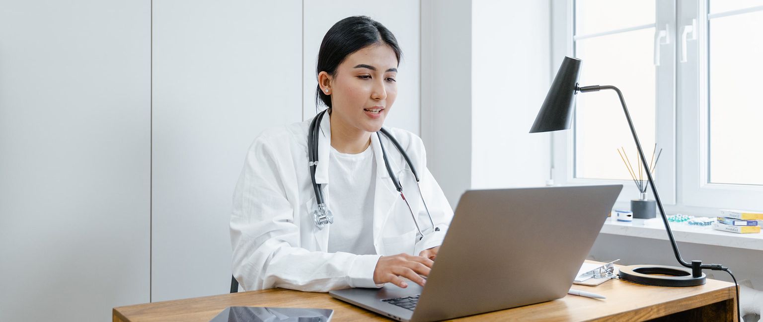 A female doctor wearing a white coat and stethoscope sits at a desk, typing on a laptop. Several items including a lamp and medications are also visible on and around the desk.