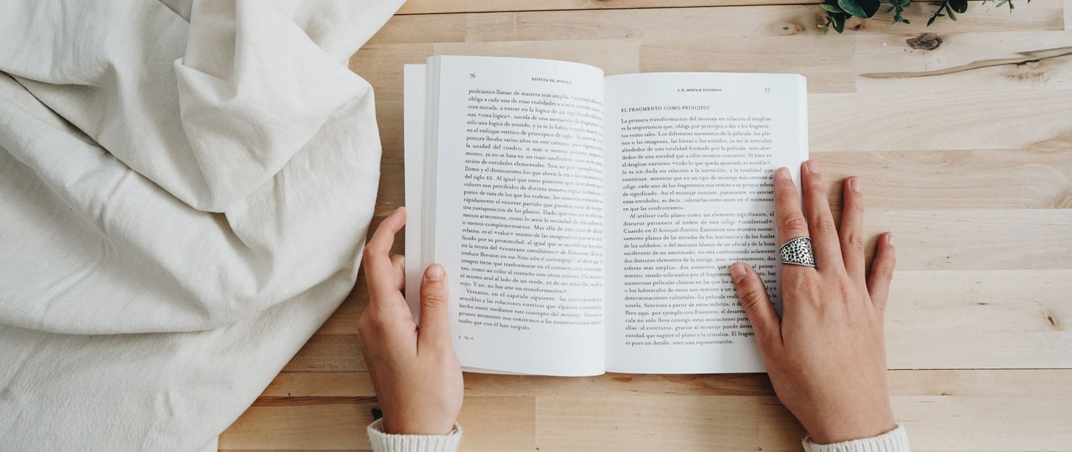 A detail shot of someone holding open a book and reading it, with their hands visible and a ring with a textured pattern on one finger.