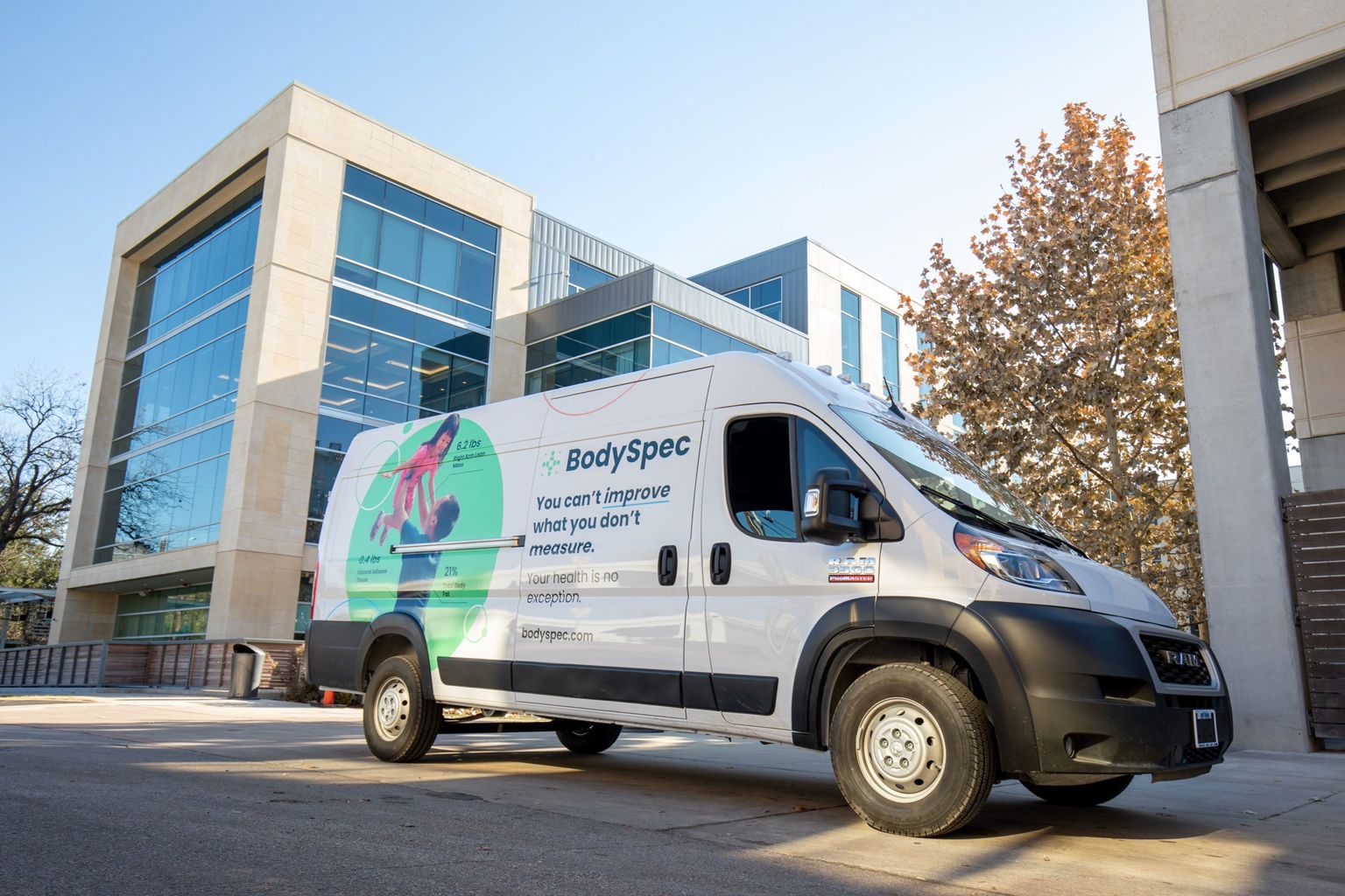 A white BodySpec Sprinter van is parked on a paved driveway in front of a modern office building with large glass windows. The van has colorful graphics with the BodySpec logo, text, and an image of a person exercising.