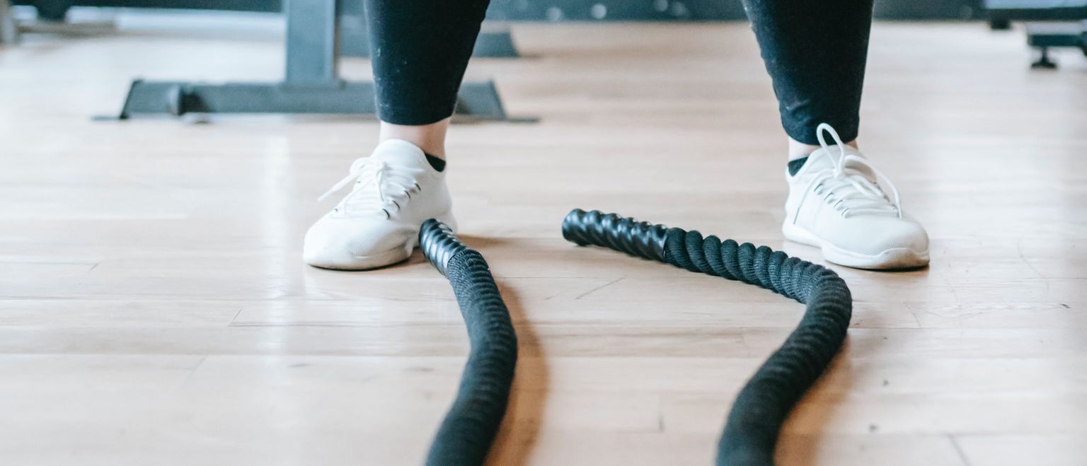 Close up of a person's feet in white sneakers standing over black battle ropes on a wooden gym floor. The person is wearing black leggings.