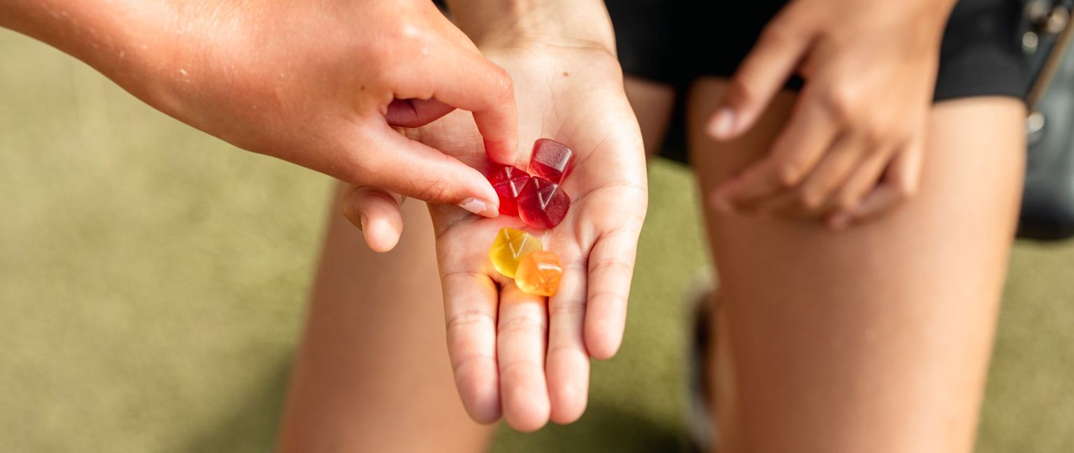 Close-up of a hand holding several square-shaped red and yellow gummies, with another hand reaching to take one.