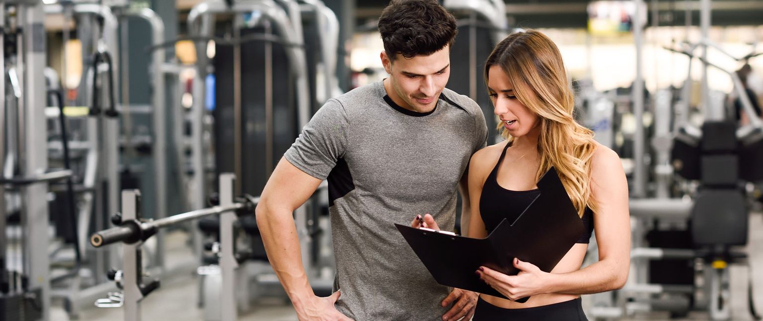 A personal trainer and his client look at a form on a clipboard in a gym.