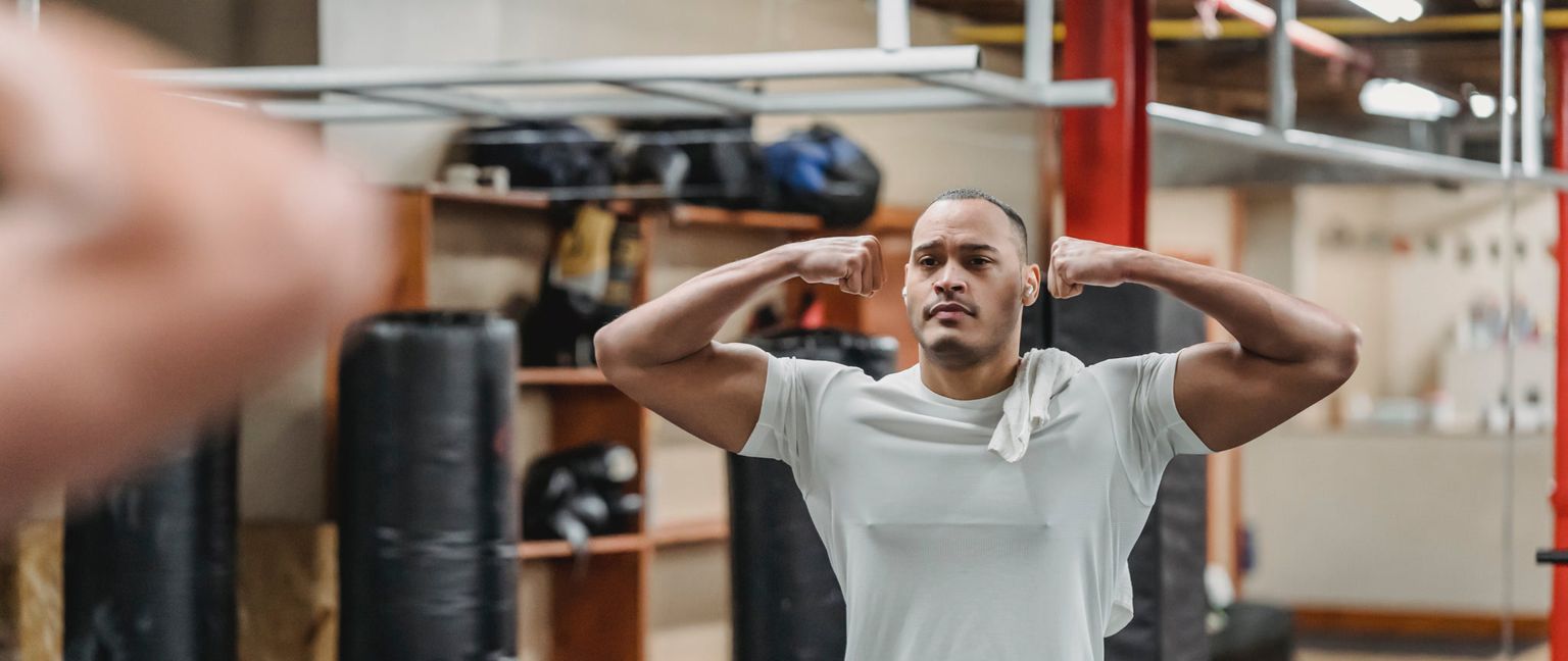 A man wearing a white t-shirt stands in a gym, flexing his biceps in a mirror reflection.