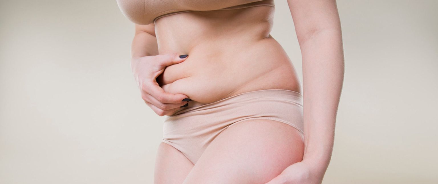 A woman wearing a nude bra and underwear pinches the skin on her stomach.