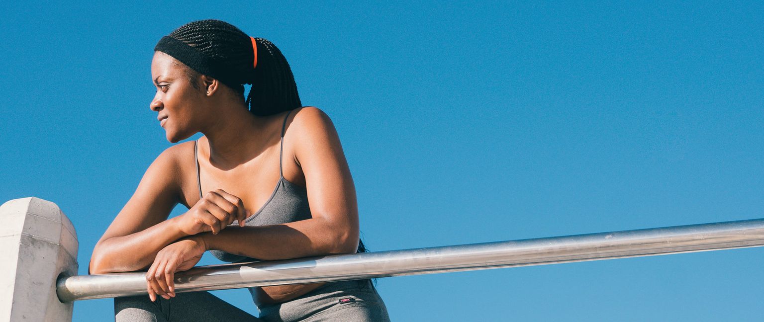 A woman wearing athletic clothing rests her arms on a metal rail with a clear blue sky in the background.