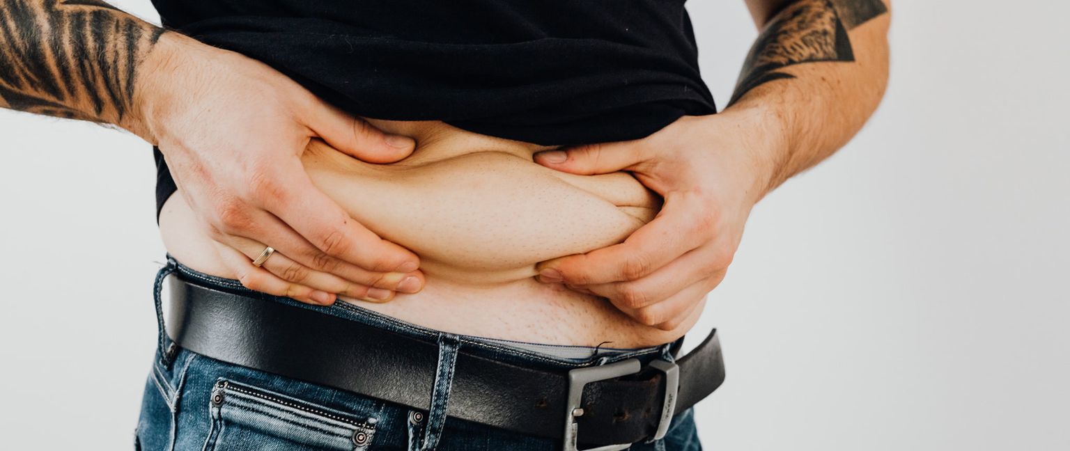 A close-up image of a man holding his belly roll with both hands while wearing a black shirt and jeans with a black belt.