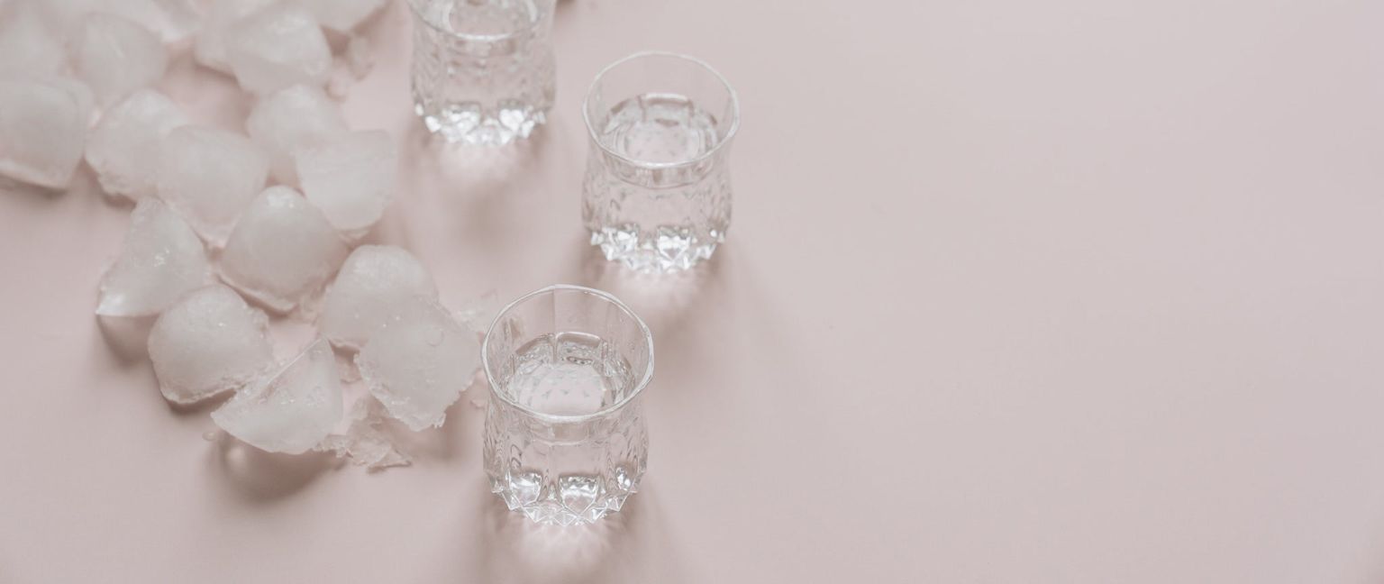 Three glasses of water sit next to a pile of ice cubes on a pink background.