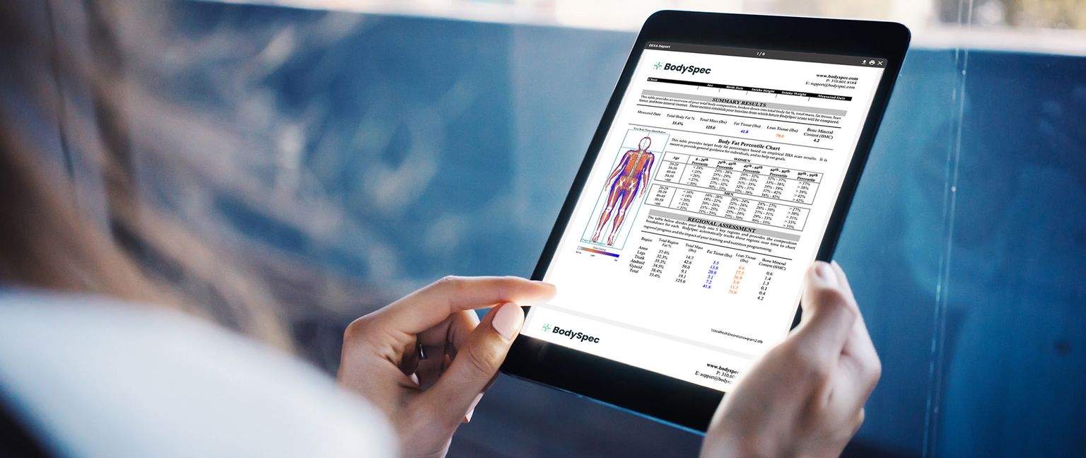 A close-up shot of a person's hand holding a tablet displaying a BodySpec DEXA report.