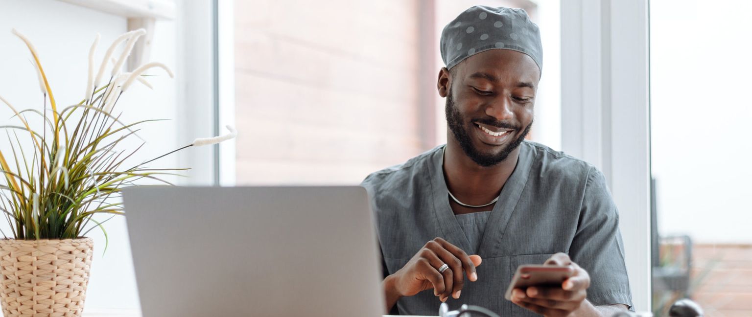 A Black man wearing a medical cap and scrubs smiles down at his phone while sitting at a desk with a laptop and plant.