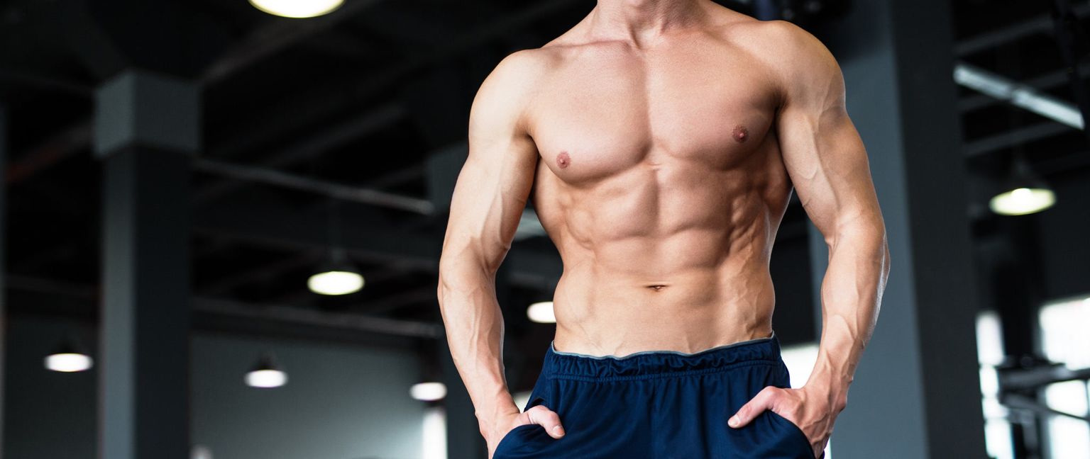 A close-up shot of a shirtless man's torso and arms, showcasing well-defined abdominal and arm muscles, standing in a gym.