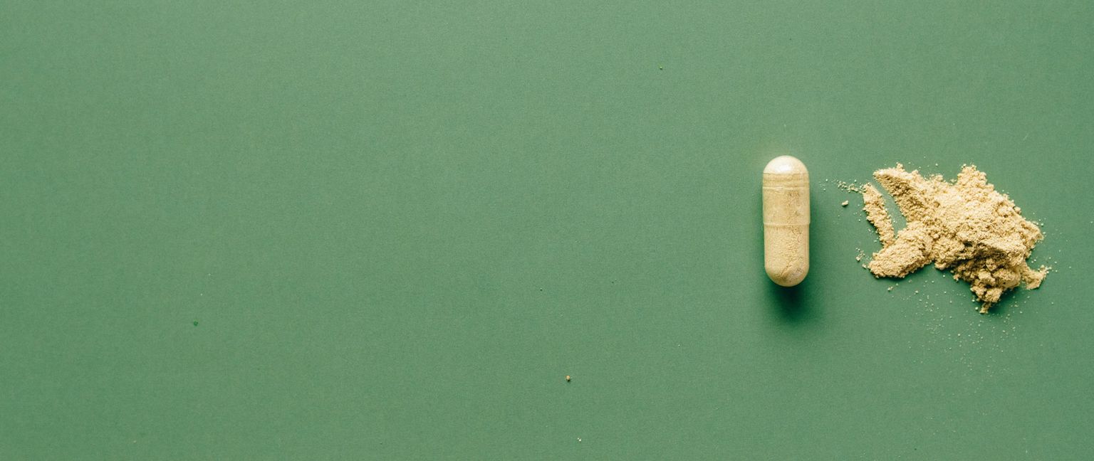 A single capsule with a small pile of beige powder next to it, all on a solid green background.